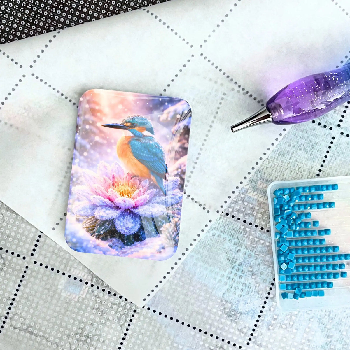 Diamond Painting Cover Minder mit Eisvogel auf Leinwand neben Glitzer-Stift und türkisen Steinen