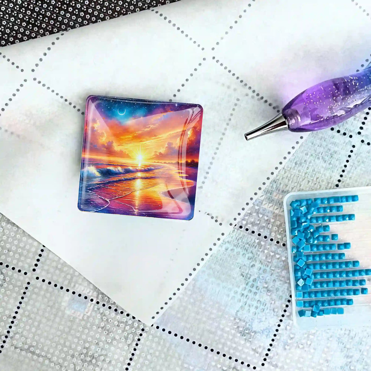 Quadratischer Diamond Painting Cover Minder mit Sonnenuntergangs-Motiv, platziert auf einer Leinwand mit Stift und Steinen
