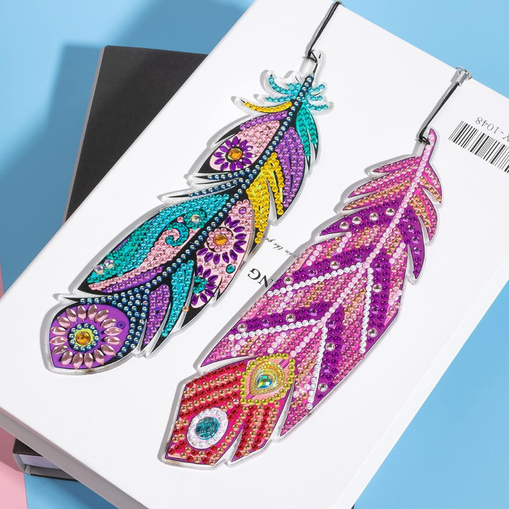 Zwei glitzernde Diamond Painting Lesezeichen im Design bunte Federn auf einem Buch liegend.