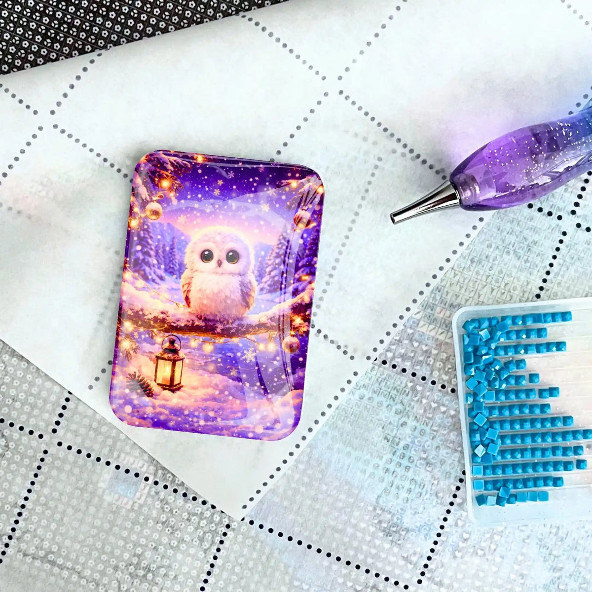 Diamond Painting Cover Minder mit Eule im Schnee auf Leinwand neben lilafarbenem Stift und Steinen