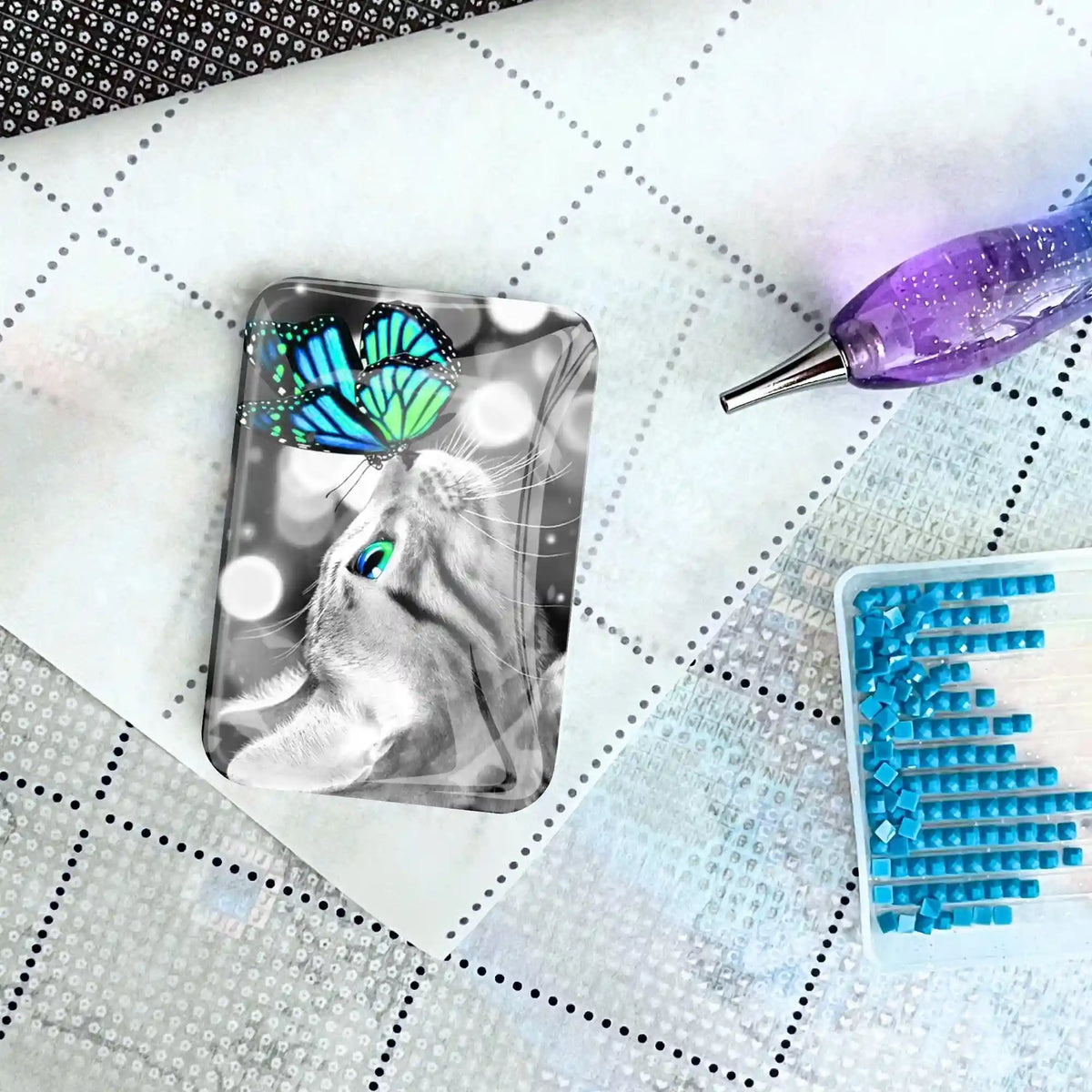 Diamond Painting Cover Minder mit Katzen-Motiv und blauem Schmetterling auf einer glitzernden Arbeitsfläche
