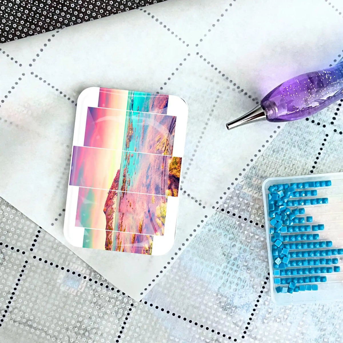 Glas Cover Minder für Diamond Painting mit Meer-Motiv auf Leinwand, daneben Stift und Schüttchen mit blauen Steinen.