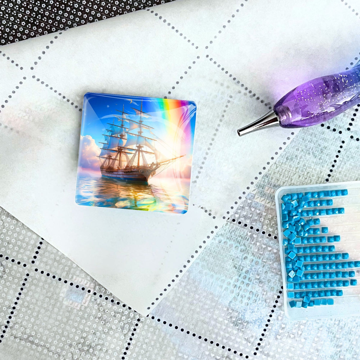 Quadratischer Diamond Painting Cover Minder mit Segelschiff-Motiv auf Leinwand, daneben Stift und blaue Steinchen.