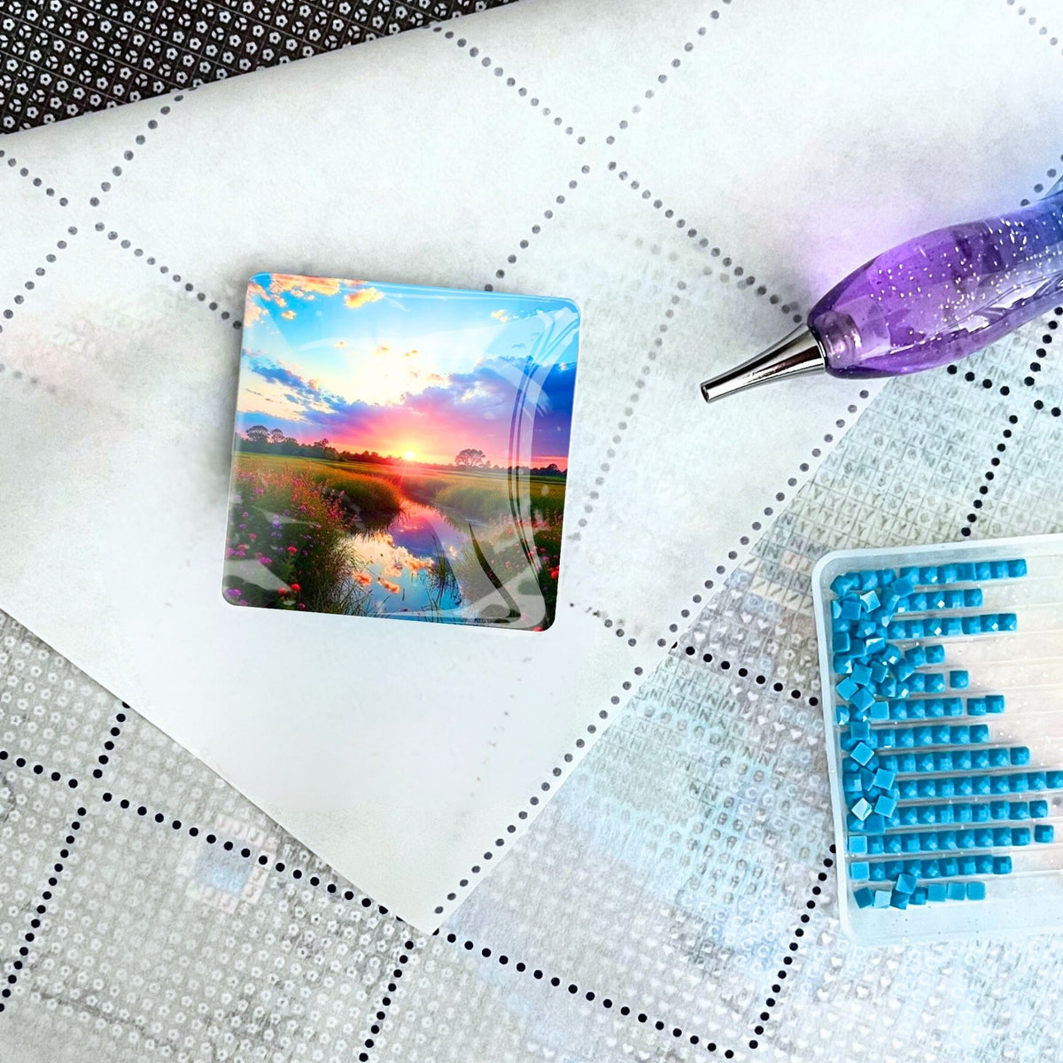 Quadratischer Diamond Painting Cover Minder Morgenrot mit Magnet auf einer Leinwand neben Stift und Diamanten-Schütteldose
