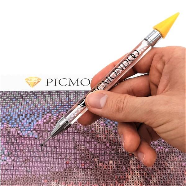 Hand hält glitzernden Picmondoo Diamond Painting Stift beim Platzieren von Steinen auf einer bedruckten Raster-Leinwand.